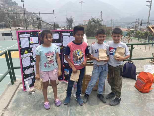 Gruppe von Kindern mit Papiertüten auf einem Basketballfeld mit Tüten und Gegenständen auf dem Boden, ein Schild mit Text links und Hintergrund-Elemente einschließlich Zaun, Pfosten, Straßenlaternen, Bäume, Gebäude, Hügel und Himmel.