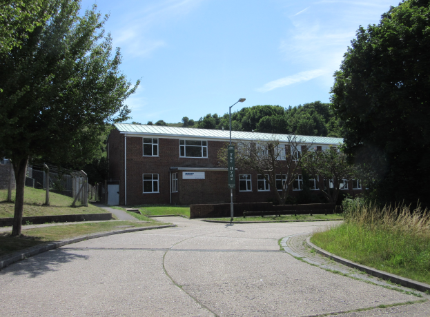 Ein Schulgebäude mit Fenstern, einem Laternenpfahl, einem Schild, einer Baumgruppe, Gras, einem Zaun und einem bewölkten Himmel, das in der Mitte eines Parks umgeben von saftigem Grün liegt.