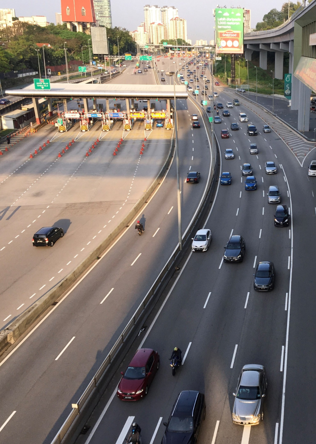Eine vielbefahrene Autobahn mit zahlreichen Autos, Motorrädern, Absperrgittern, Schildern, Laternenmasten, einer Brücke, Bäumen, Gebäuden und einem klaren blauen Himmel.