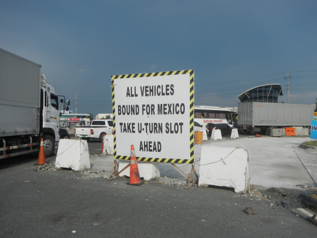 Ein Lkw parkt auf einem Parkplatz neben einem Schild mit der Aufschrift "Alle Fahrzeuge nach Mexiko gebunden". Es sind Verkehrskegel, Bretter und andere Gegenstände verstreut und im Hintergrund sind Gebäude, Bäume, Pfosten und ein klarer blauer Himmel zu sehen.