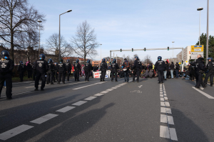 Eine Gruppe von Polizisten in schwarzen Uniformen und Helmen, die auf der Seite einer Straße mit Laternenmasten, Ampeln, Bäumen, Gebäuden und einem klaren blauen Himmel im Hintergrund stehen.
