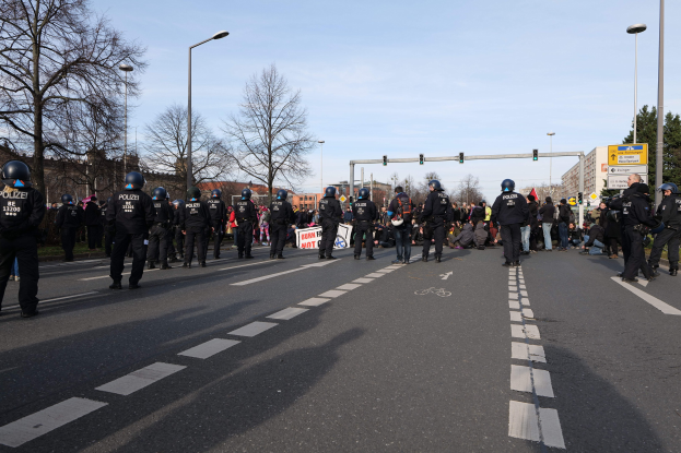 Eine Gruppe von Polizisten in schwarzen Uniformen und Helmen, die auf der Seite einer Straße mit Laternenmasten, Ampeln, Bäumen, Gebäuden und einem klaren blauen Himmel im Hintergrund stehen.