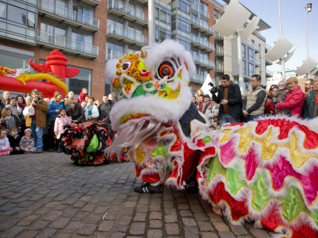 Eine Löwen-Tanzvorstellung während eines chinesischen Neujahrsfestes in Amsterdam, mit einer Zuschauermenge, einige fotografieren das Ereignis, vor einem Hintergrund von Gebäuden, Laternenmasten und einem klaren blauen Himmel.