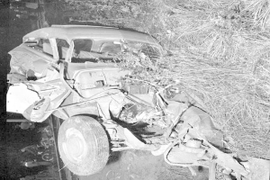 Schwarz-weiß-Foto eines schwer beschädigten Autos mit eingedellter Front und zersplitterten Fenstern am Straßenrand, mit einem Baum und verstreuten Gegenständen im Hintergrund.