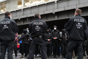 Eine Gruppe von Polizisten In Uniform Steht Vor Einer Menge Von Menschen, Die Schwarze Uniformen Und Masken Tragen, Mit Einer Stadtbrücke Und Einem Gebäude Im Hintergrund, Während Eines Protests.