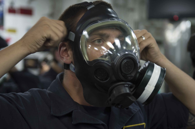 Ein Mann mit einer Gasmaske in einer Fabrik, umgeben von ein paar anderen Menschen, mit einem unscharfen Hintergrund.