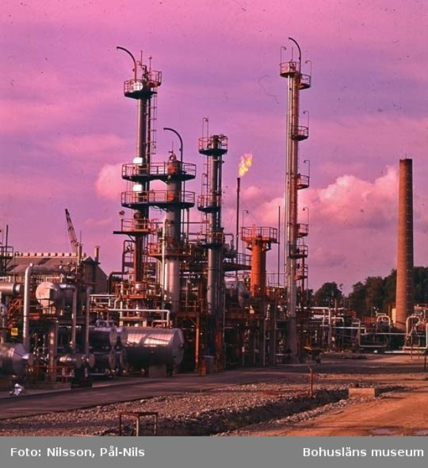 Ölraffinerie mit ausgedehntem Netzwerk von Rohren, Ventilen, Metallstrukturen, Containern, Bäumen, einem Gebäude, einem Kran und einem bewölkten Himmel, mit Text unten.