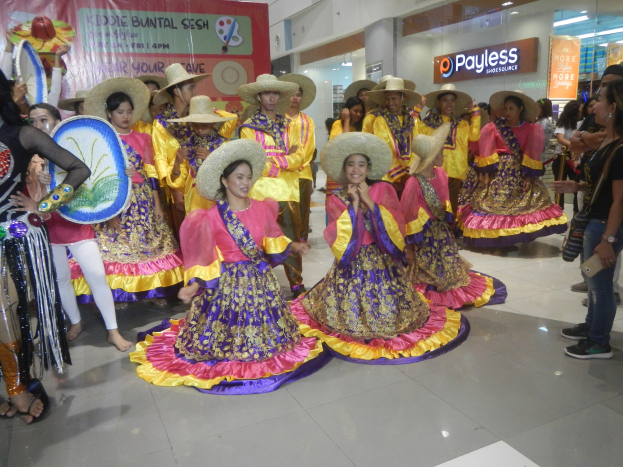Eine Gruppe von Menschen in bunten Kleidern und Hüten, die in einem Einkaufszentrum während eines festlichen Ereignisses tanzen, während andere Musikinstrumente halten und ein Banner im Hintergrund zu sehen ist.