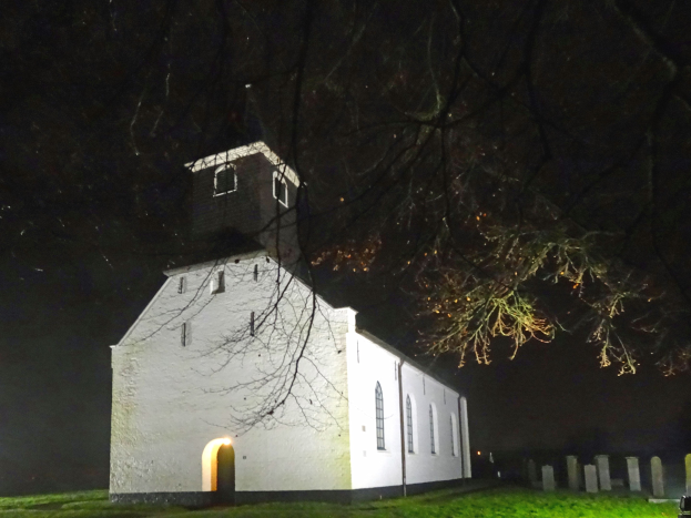 Weiße Kirche mit einem beleuchteten Kirchturm in der Nacht, umgeben von Gras und einem Baum im Vordergrund, mit Gräbern und einem dunklen Himmel im Hintergrund.