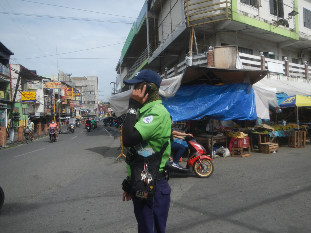 Ein Polizeibeamter in grüner und blauer Uniform und einer blauen Mütze steht an der Seite einer Straße und spricht in ein Handy, während Fahrzeuge auf der Straße hinter ihm fahren, Stände mit verschiedenen Gegenständen rechts von ihm und ein bewölkter Himmel mit einem Gebäude mit Fenstern im Hintergrund.