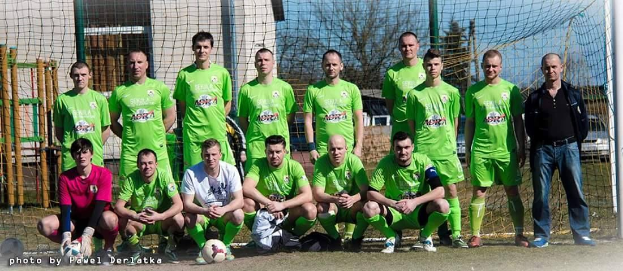 Eine Gruppe von Männern in grünen T-Shirts steht auf einem Fussballfeld, einige hocken, mit einem Ball und einem Torpfosten im Hintergrund, sowie Bäumen und Gebäuden.