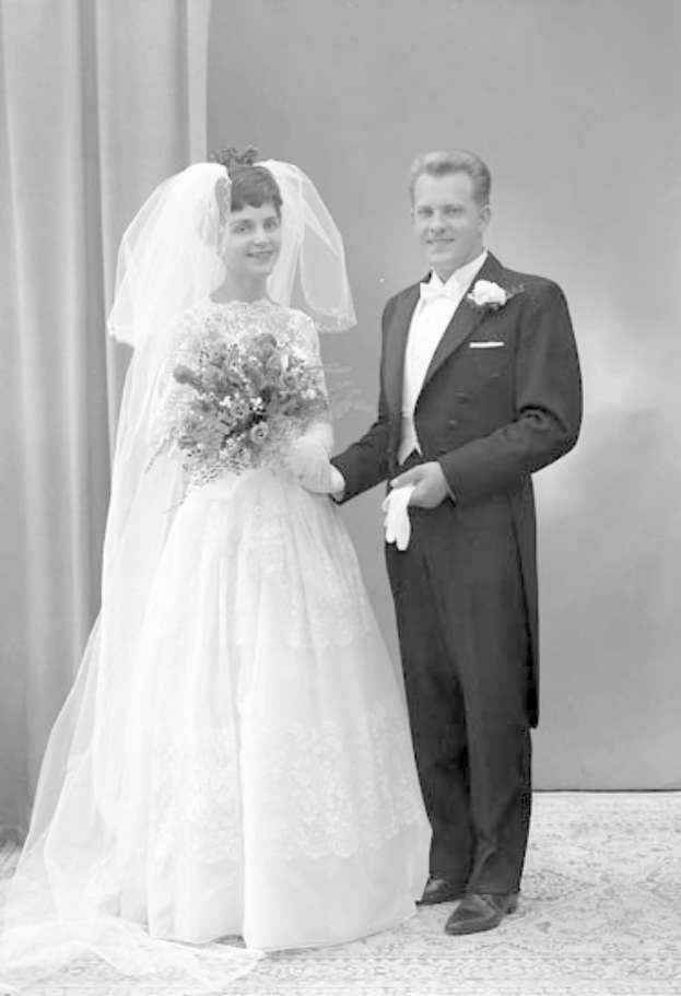 Schwarzes und weißes Foto eines lachenden Mannes und einer Frau in Hochzeitskleidung, die gemeinsam stehen, die Frau hält einen Strauß, mit einem Vorhang und einer Wand im Hintergrund.