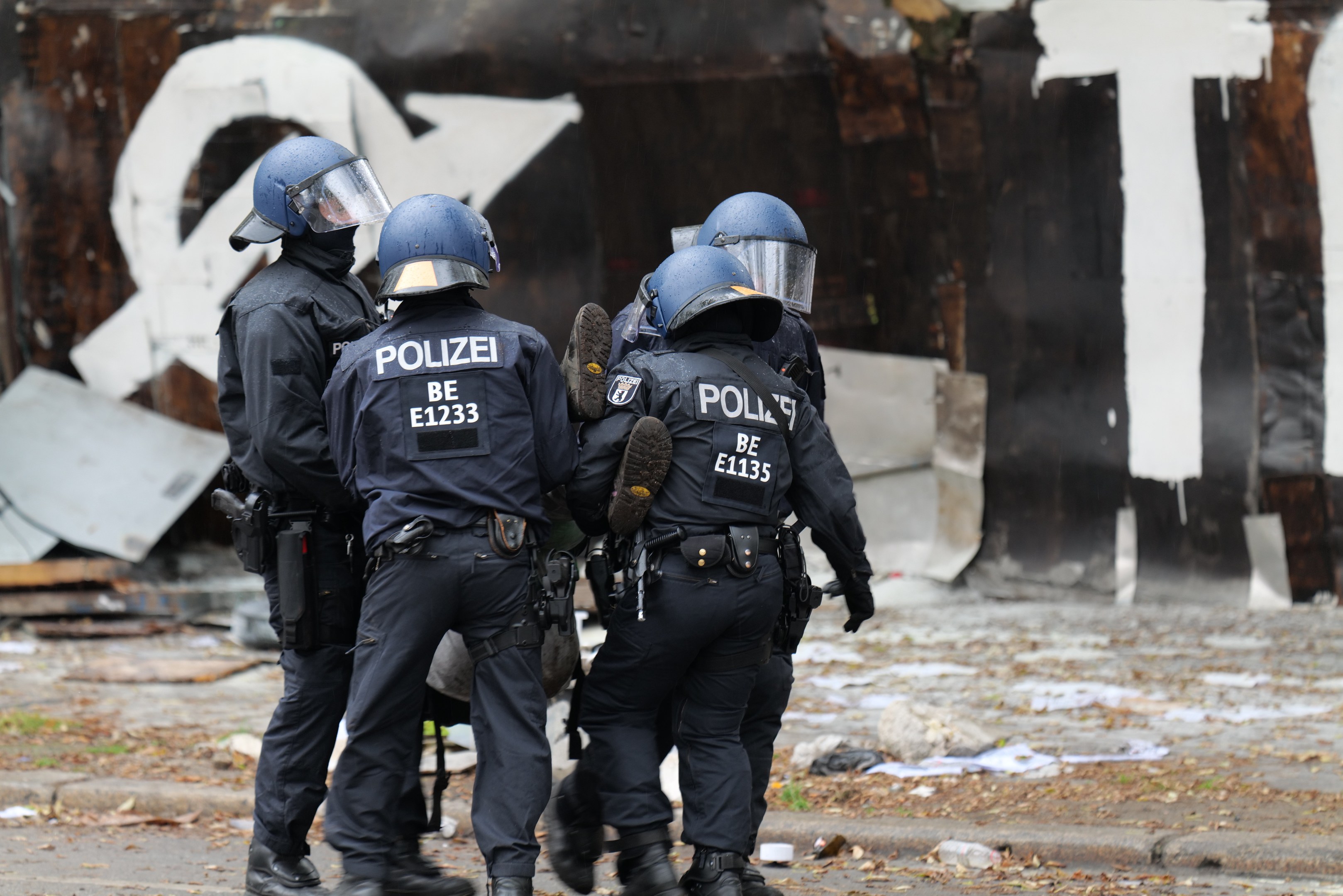 Polizeibeamte in schwarzen Uniformen und blauen Helmen stehen vor einem mit Graffiti bedeckten Gebäude während einer Unruhen, mit verstreuten Papieren und Gegenständen auf dem Boden.