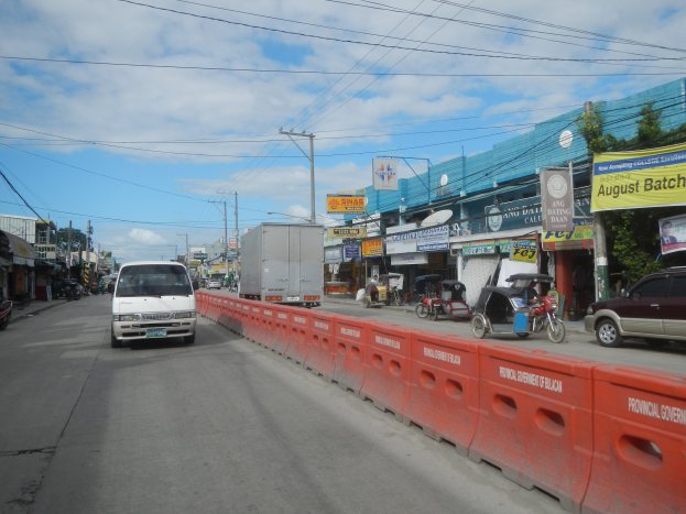 Eine belebte Stadtstraße mit mehreren Fahrzeugen, einer Mittelinsel, Gebäuden mit Schildern, Strommasten mit Drähten, Bäumen und einem bewölkten Himmel.