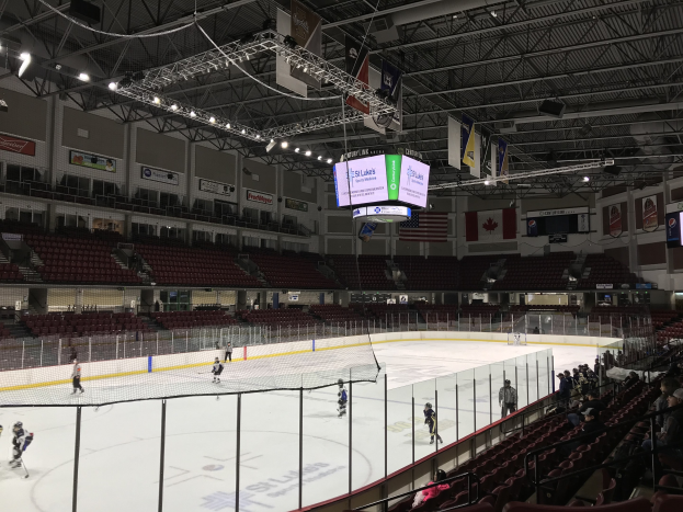 Eishockeyspiel in einer großen Arena mit einem zentralen Netz, Zuschauern auf Stühlen, Deckenbeleuchtung und Trägern sowie Bannern an den Wänden, die 'St. Louis Blues Arena' beschriftet sind.