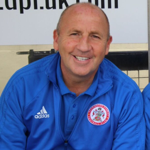 Zwei Männer in einem Raum, einer trägt eine blaue Jacke und lächelt neben einem anderen Mann, mit einer Tafel an der Wand im Hintergrund.