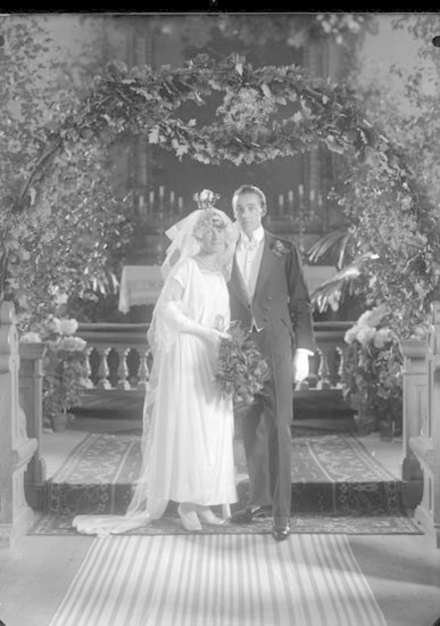 Ein Mann und eine Frau stehen vor einem mit Pflanzen und Blumen geschmückten Hochzeitsbogen, die Frau hält einen Strauß, auf einem Teppichboden, in Schwarz-Weiß dargestellt.