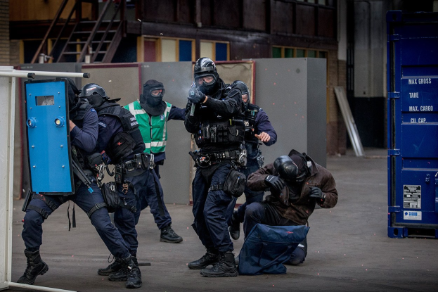 Polizeibeamte in Einsatzausrüstung stehen vor einem Gebäude in einer Reihe, wobei eine Person im Vordergrund liegt und ein blaues Objekt mit Text auf der rechten Seite zu sehen ist.