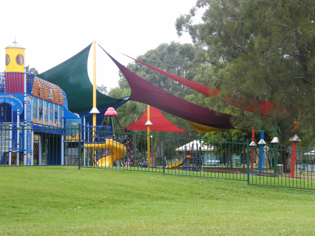 Ein Park mit einem Spielplatz, der von einem Metallzaun umgeben ist, mit Kindern, die auf Rutschen, Stangen und einem Zelt spielen, umgeben von grünem Gras, Bäumen und einem klaren blauen Himmel.