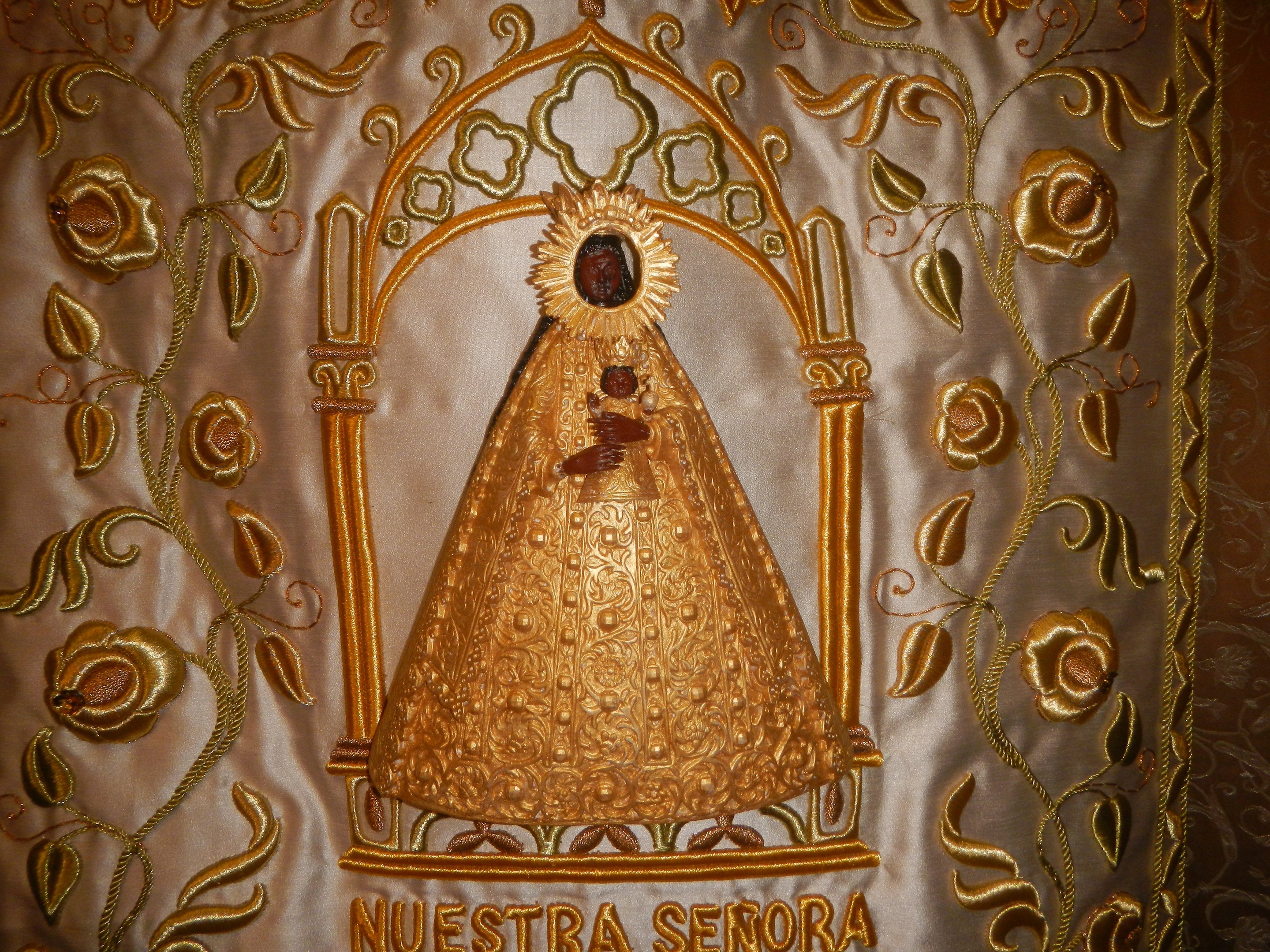 Goldbrokat-Chorrock mit einer serenitären Frau in einem hochgeschlossenen, langärmligen Kleid mit einem Dutt, umgeben von detailreichen Designs, Texten und goldenen Bordüren.