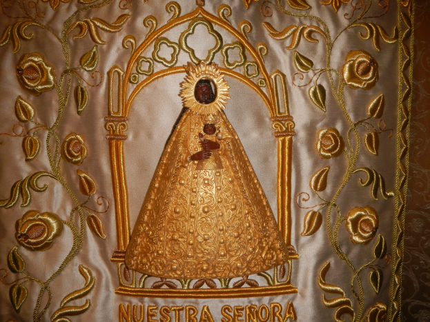 Goldbrokat-Chorrock mit einer serenitären Frau in einem hochgeschlossenen, langärmligen Kleid mit einem Dutt, umgeben von detailreichen Designs, Texten und goldenen Bordüren.