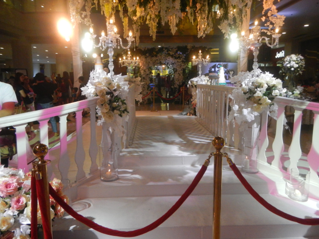 Eine Hochzeitszeremonie in einem Einkaufszentrum mit einem weißen Teppichboden, Geländern, Pfosten mit Seilen, Blumenbouquets, Kronleuchtern mit Blumen, Lichtern und anderen dekorativen Elementen, mit vielen Menschen, einige mit Taschen, und einer Decke, die mit Lichtern geschmückt ist.