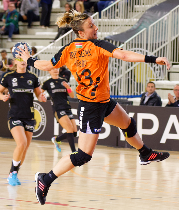 Eine Frau in einem orangen T-Shirt und schwarzen Shorts spielt Handball mit einem Ball in der Hand, mit Zuschauern und einem Banner mit der Aufschrift "Krp Slezak - Handball-Weltmeisterschaft 2019" im Hintergrund.