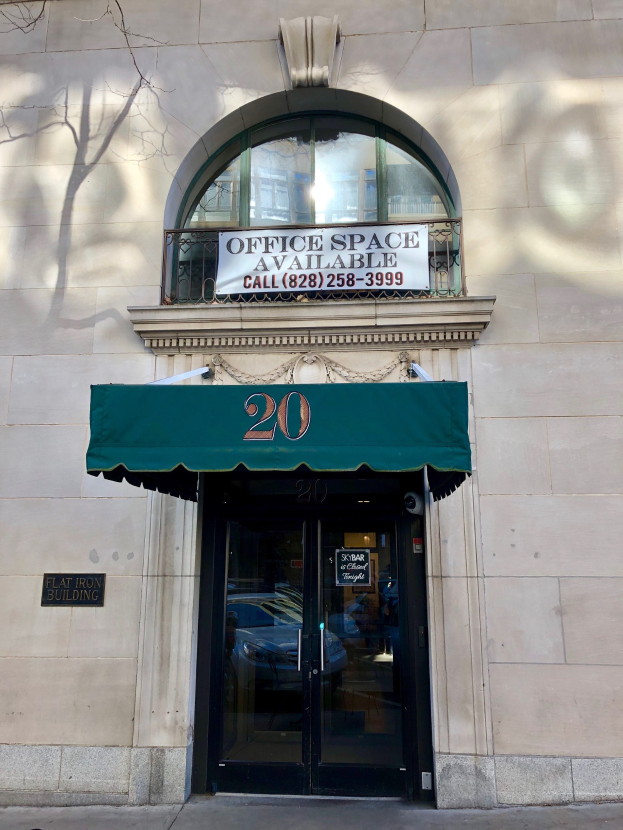 Ein Gebäude mit einer Glastür, einer grünen Markise, einem Schild mit der Aufschrift "Büroräume zu vermieten", einem Geländer und einem Baum links.