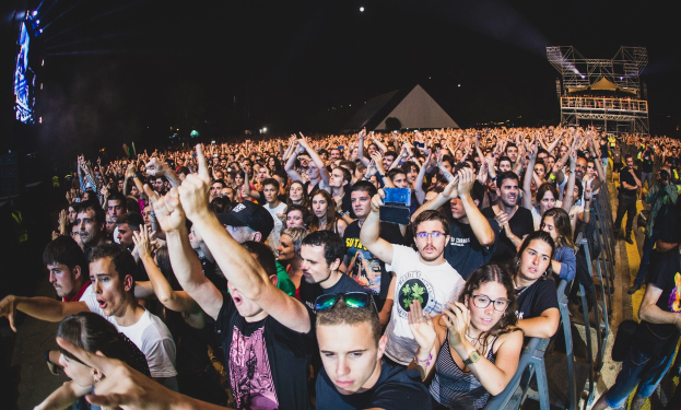 Menge auf einem Musikfestival mit erhobenen Händen, einige sitzend und andere stehend, in der Nähe eines Metallzauns mit Gebäuden und Lichtern im Hintergrund.