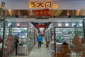 Innenraum einer chinesischen Apotheke mit Glaswänden, Regalen mit verschiedenen Artikeln, verstreuten Kartons auf dem Boden, einer Pflanze auf der linken Seite und einem Namensschild oben.