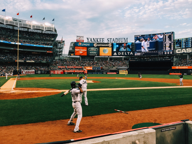 Baseballspiel im Yankee Stadium mit Spielern auf dem Feld und Zuschauern in den Rängen, umgeben von Stadionmerkmalen unter einem bewölkten Himmel.