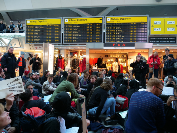 Große Gruppe von Menschen in einem Flughafen, einige sitzen mit Taschen und Papieren, andere stehen, mit Informationsschildern, Schaufensterpuppen in Kleidern und Deckenbeleuchtung, was auf eine Protestaktion hindeutet.