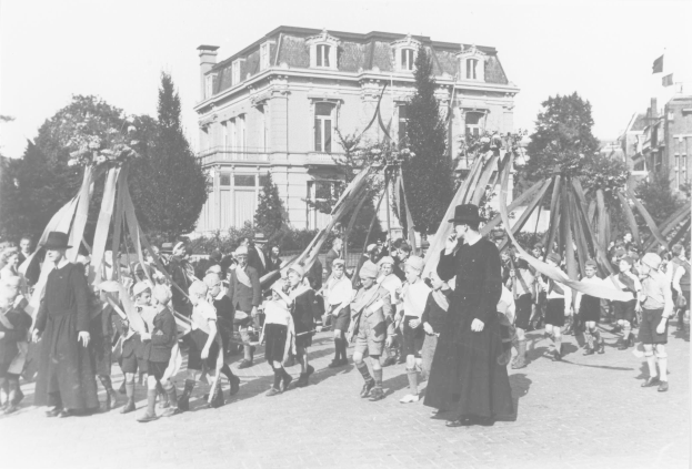 Schwarz-weißes Bild einer Gruppe von Menschen, die eine Straße entlanggehen, einige halten Fahnen, mit Hüten, Bäumen, Gebäuden und einem Maibaum im Hintergrund.