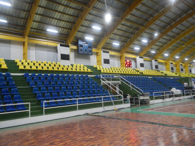 Große Hallensportanlage mit Holzboden, blauen und gelben Sitzplätzen, Geländern, Stufen zum Sitzbereich, wandmontierten Bildschirmen, Deckenleuchten und einigen Personen.