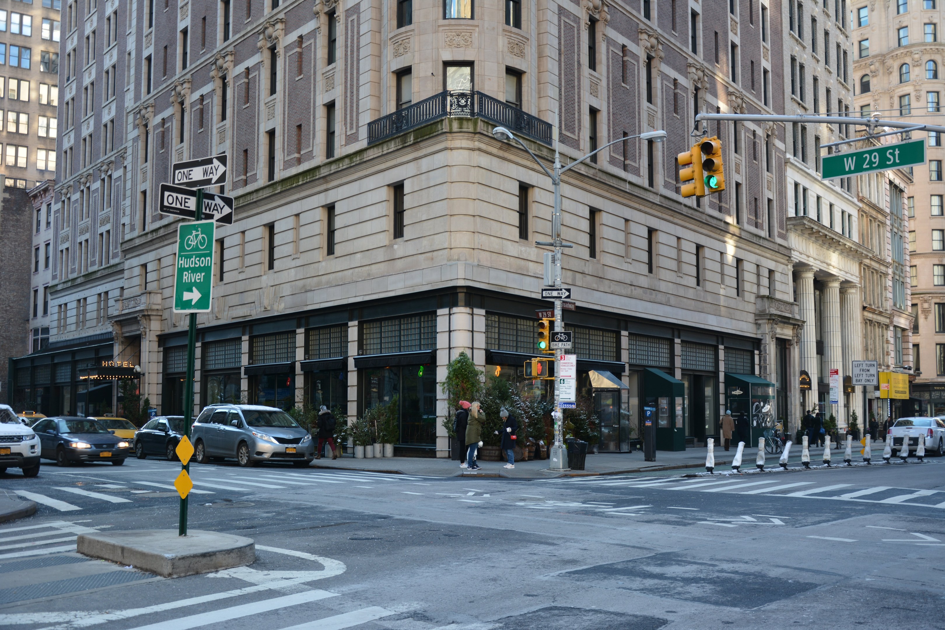 Eine belebte Stadtstraße mit hohen Gebäuden, Fahrzeugen, Verkehrsampeln, Schildern, Fußgängern auf dem Gehweg und einem geparkten Fahrzeug.
