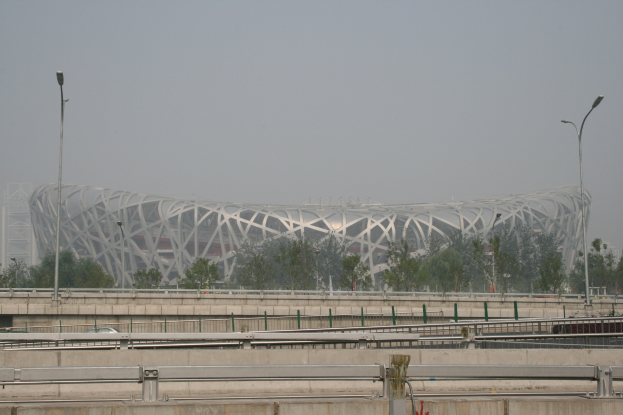Olympiastadion in Peking, China, mit Brücke, Geländern, Pfählen, Lichtern, Bäumen und einem Gebäude vor einem klaren blauen Himmel.