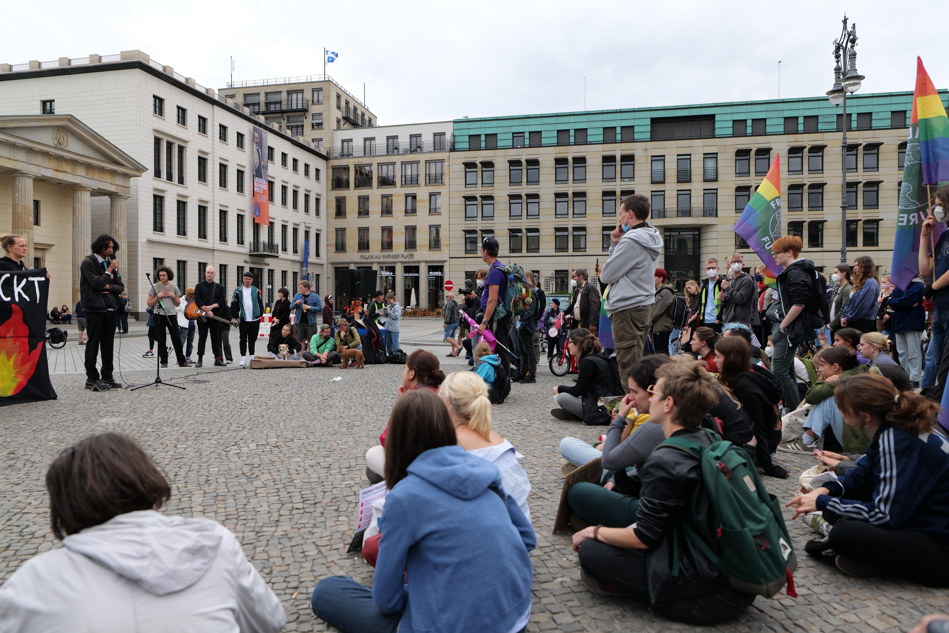 Eine Gruppe von Menschen, die auf dem Boden vor einer Menge sitzt, die Fahnen und Plakate hölt, mit einer Person, die in ein Mikrofon spricht, einer Statue und Gebäuden im Hintergrund während einer anti-schwulen Demonstration in Berlin, Deutschland.