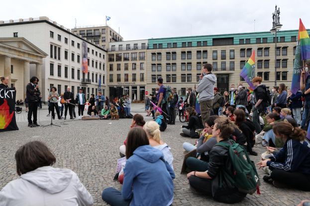 Eine Gruppe von Menschen, die auf dem Boden vor einer Menge sitzt, die Fahnen und Plakate hölt, mit einer Person, die in ein Mikrofon spricht, einer Statue und Gebäuden im Hintergrund während einer anti-schwulen Demonstration in Berlin, Deutschland.