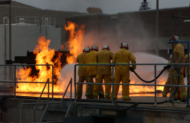 Eine Gruppe von Feuerwehrleuten in Helmen, die Rohre halten, steht auf dem Dach eines Gebäudes mit Geländern und Treppen, die unten sichtbar sind, mit einem weiteren Gebäude und dem Himmel im Hintergrund.