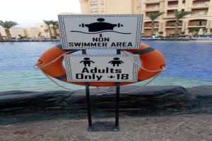 Ein Schwimmbad mit einem Schild "Nur für Erwachsene, die nicht schwimmen können" im Vordergrund, umgeben von Bäumen und Gebäuden unter einem klaren blauen Himmel, mit einem Rettungsring in der Mitte des Pools.