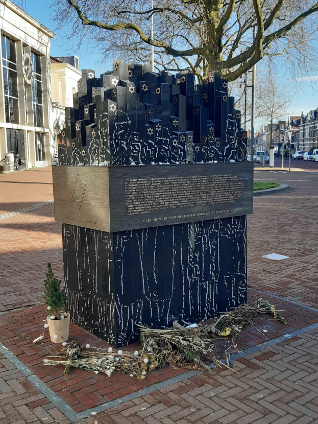 Denkmal für Holocaust-Opfer in einem Stadtplatz mit einem Baum, Topfpflanze, verstreuten trockenen Blättern und städtischem Hintergrund einschließlich Gebäuden, Fahrzeugen, Pfählen und Bäumen.