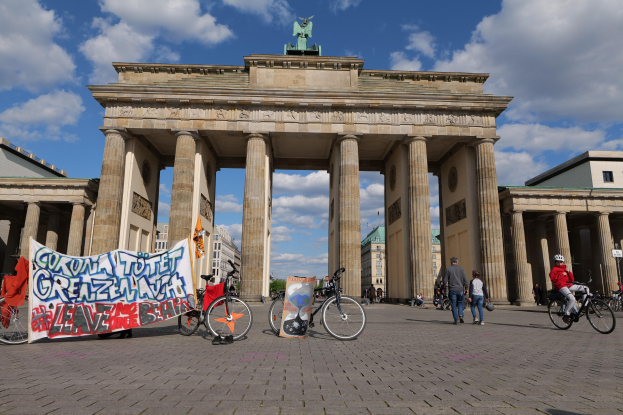 Gruppe von Menschen, die Fahrräder vor dem Brandenburger Tor fahren, einem prächtigen Bogen mit Säulen und einer Quadriga-Statue darauf, mit einem Banner und Gebäuden im Hintergrund.