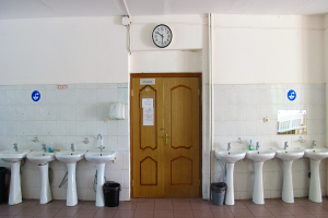 Reihe von Waschbecken mit Wasserhähnen, Mülltonnen auf dem Boden, einer Tür im Hintergrund, einer Uhr an der Wand und einem Spiegel auf der rechten Seite.