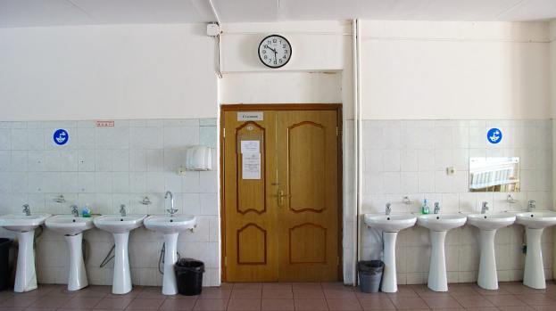 Reihe von Waschbecken mit Wasserhähnen, Mülltonnen auf dem Boden, einer Tür im Hintergrund, einer Uhr an der Wand und einem Spiegel auf der rechten Seite.