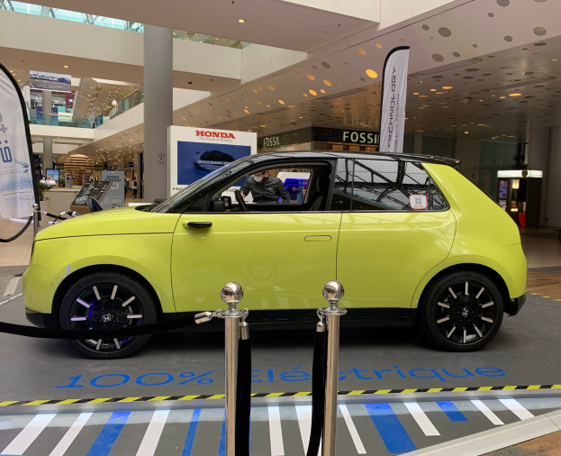 Ein gelber wasserstoffbetriebenes Elektroauto vor einem Gebäude geparkt, umgeben von Barrieren und Bannern, mit Menschen in der Nähe und einer Topfpflanze.