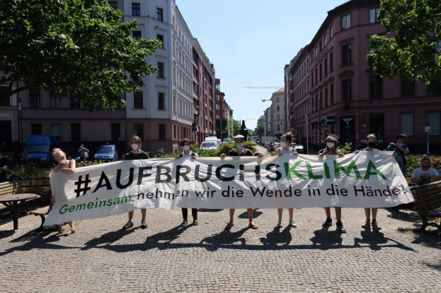 Eine Gruppe von Menschen in Masken und mit einem Banner mit der Aufschrift 'Aufbruchsklima' protestiert gegen den Klimawandel in Berlin, Deutschland, vor einem Gebäude, das von Bänken, Tischen, Pflanzen, Bäumen, Fahrzeugen, Laternenmasten, Schildern und anderen Menschen umgeben ist, mit dem Himmel im Hintergrund.