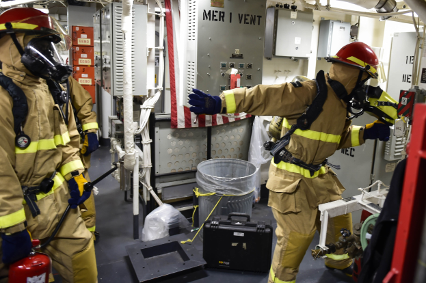 Drei Feuerwehrleute in Schutzausrüstung stehen in einem Raum mit einem Feuerlöscher links, einer Maschine rechts und verstreuten Gegenständen auf dem Boden.