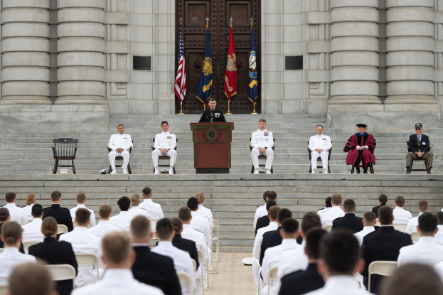 Gruppe von Menschen, die auf Stühlen vor einem Gebäude mit Säulen und einer Tür sitzen, mit einem Podium und Mikrofon in der Mitte und Flaggen im Hintergrund, was auf eine Abschlussfeier hinweist.