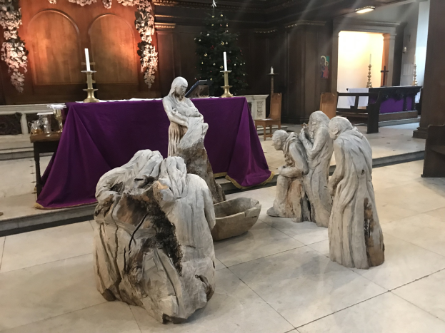 Eine Krippe in einer Kirche mit Statuen auf dem Boden, einem Tisch mit einem Tuch und Kerzenständern, Stühlen, einem geschmückten Weihnachtsbaum und einer Wand im Hintergrund.