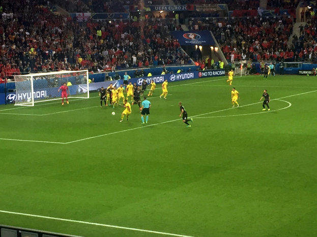 Eine Gruppe von Menschen, die auf einem Feld in einem Stadion mit einem Netz auf der linken Seite Füßball spielen, eine Menge auf den Rängen und beleuchtete Tafeln im Hintergrund.
